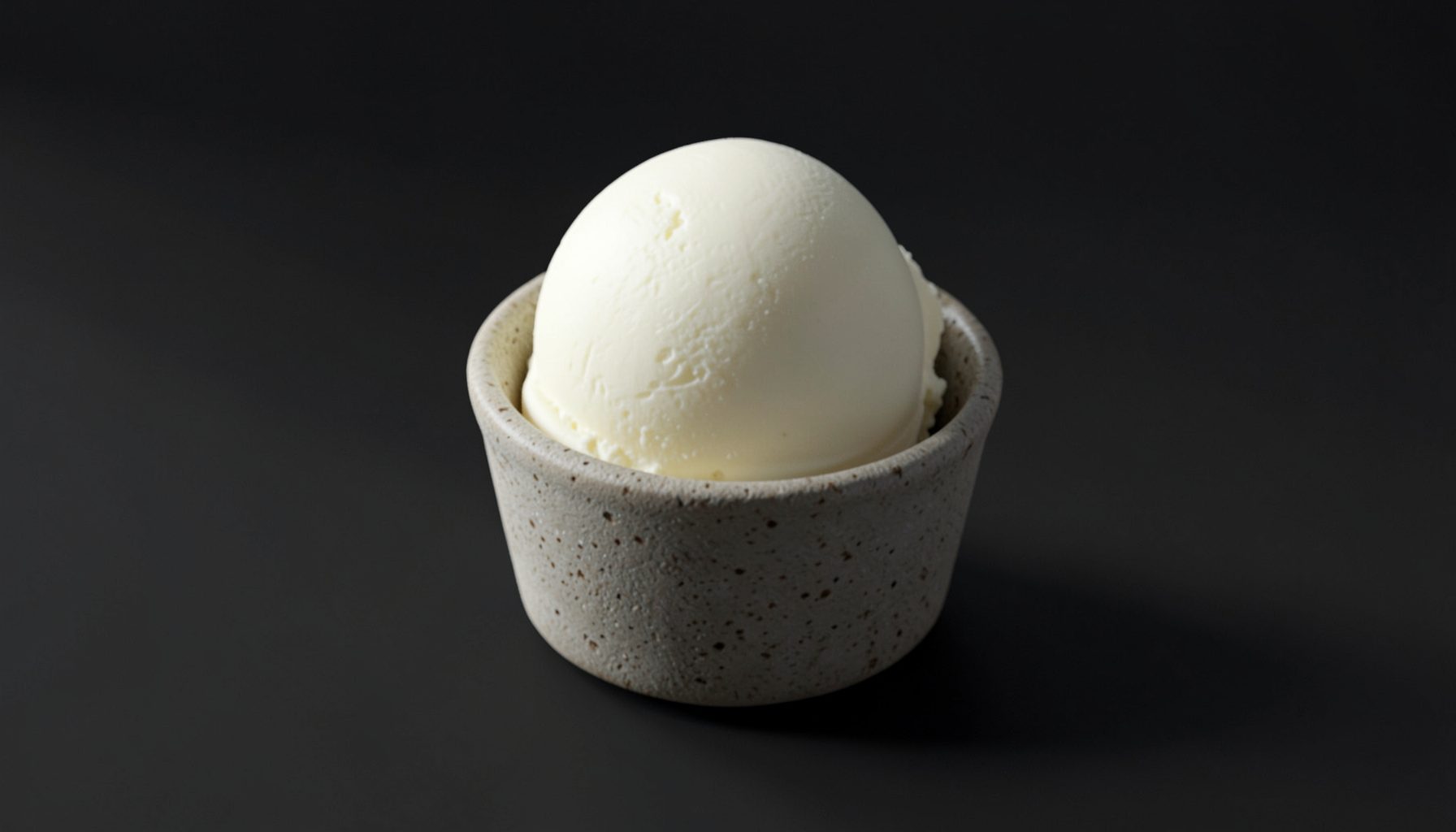 A single scoop of creamy off-white vanilla low-cal ice cream in a textured stoneware cup, soft natural light from the side, dark moody background.